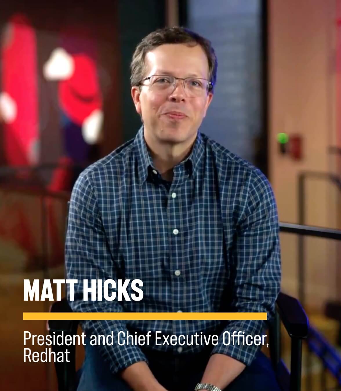 A person in a blue checkered shirt sits in a modern office setting with on‑screen text reading MATT HICKS, President and Chief Executive Officer, Redhat.