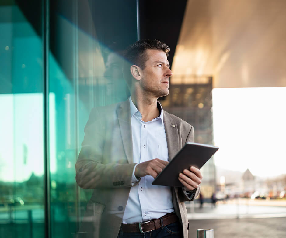 Una persona de pie frente a un edificio de vidrio mirando a la distancia con una tableta en la mano.
