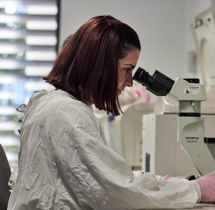 person in lab coat looking through microscope