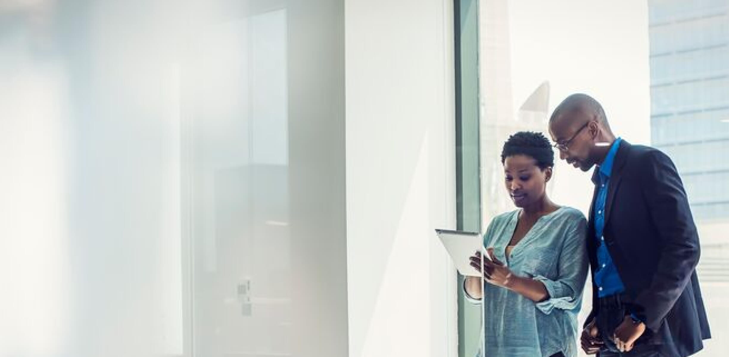 Two colleagues discussing work while reviewing a tablet near a large office window.