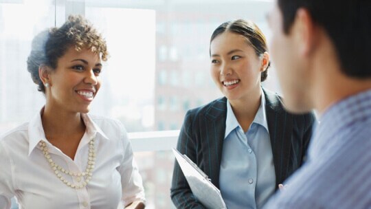 Coworkers having a discussion in a bright office with large windows and city views.