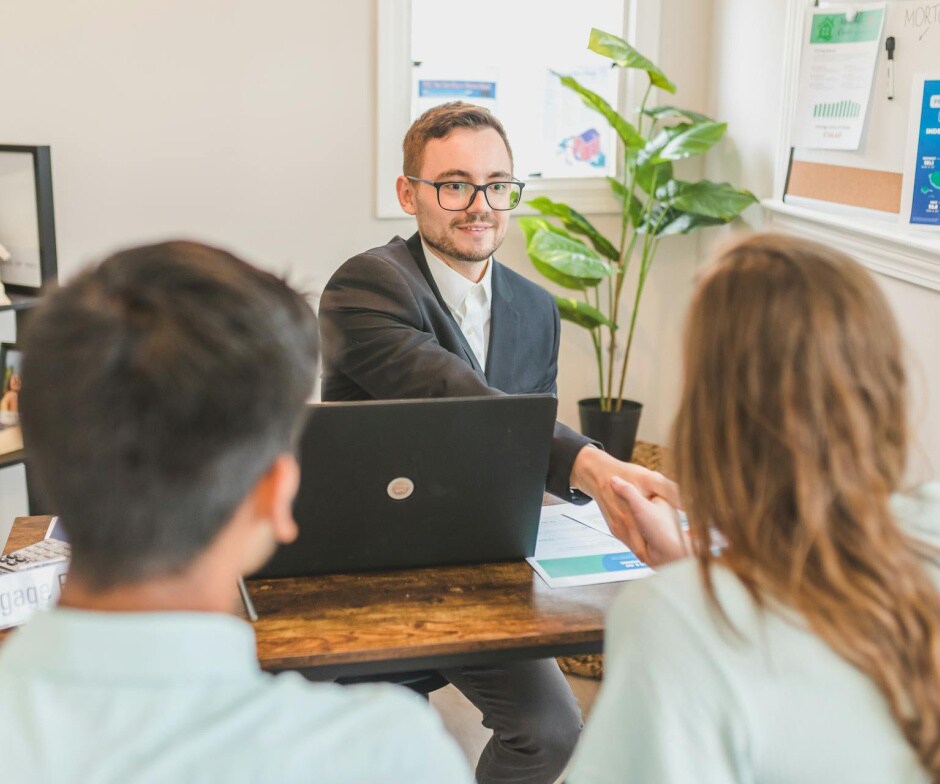 A person in business attire sits at a desk with a laptop, meeting with two individuals seated across and extending a handshake in an office setting.
