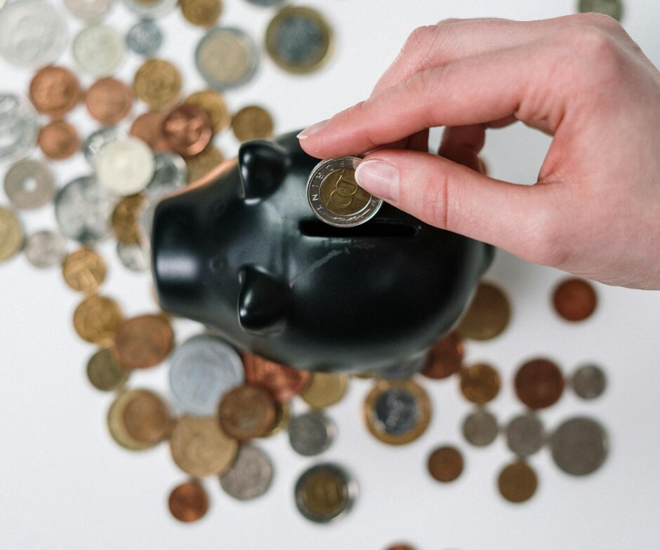 Close‑up of a hand inserting a coin into a black piggy bank with various coins spread around.