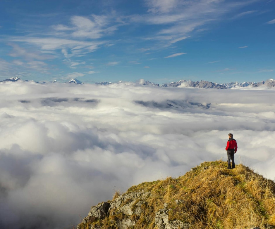 A lone hiker stands on a grassy mountain peak above a vast sea of clouds with distant snow-covered mountains under a blue sky.