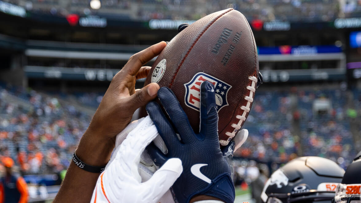 Two football players’ hands reaching up to catch an NFL football during a game.