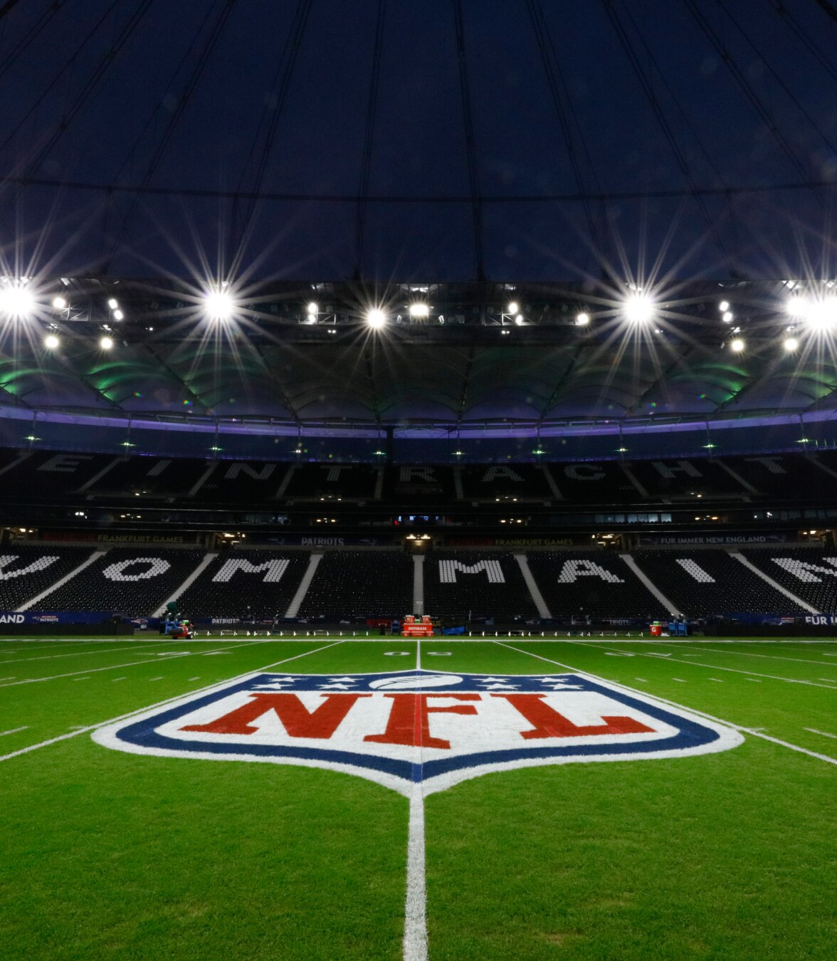 Nighttime view of an illuminated NFL football field with the NFL logo at midfield inside a large stadium.