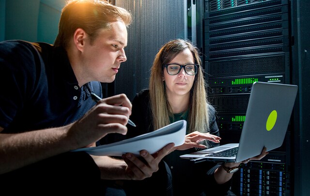 one person looking at laptop and the other person taking notes while looking at laptop in a server room
